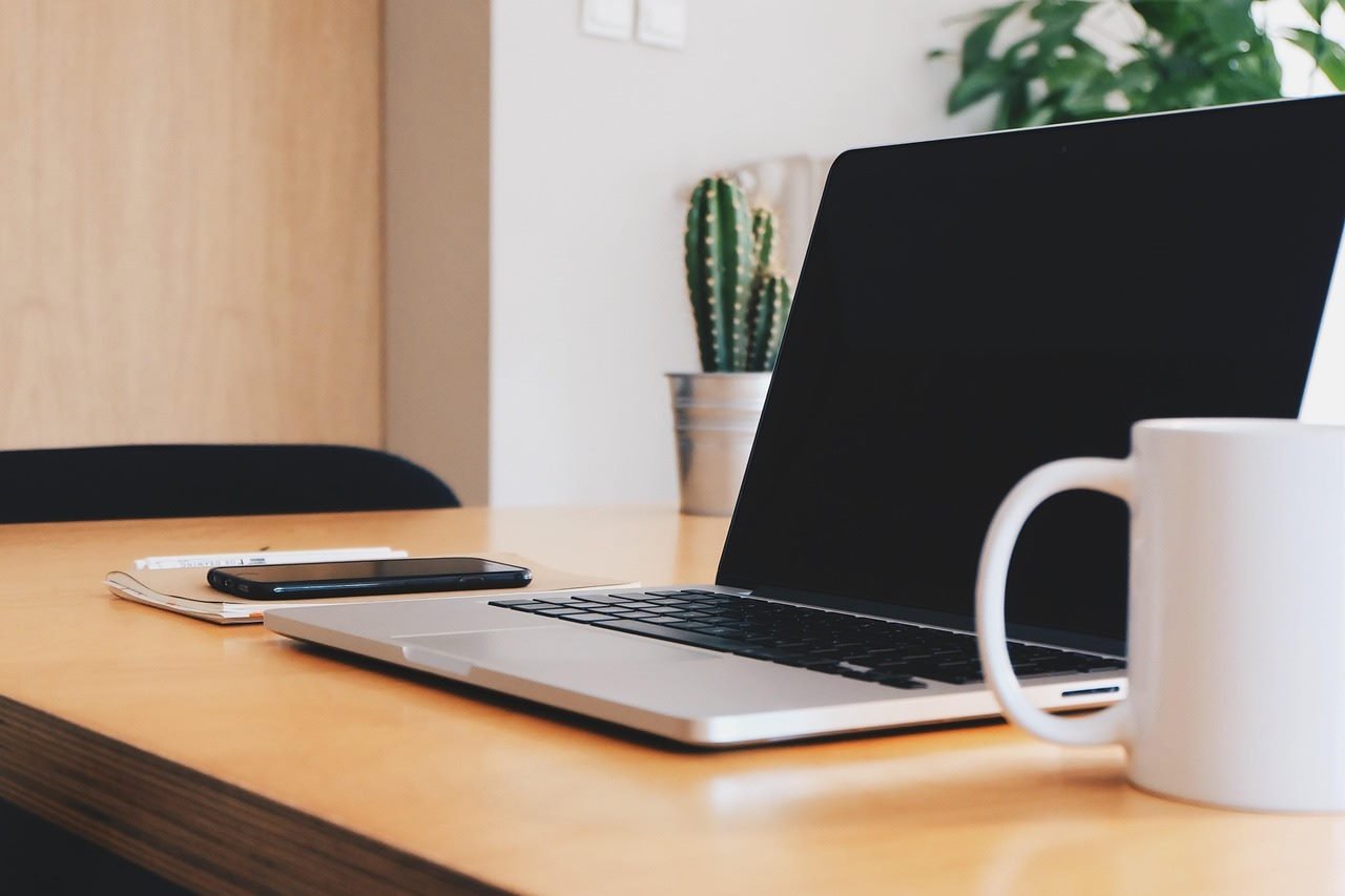 A workspace image - with a laptop, coffee cup, phone, notepad, and a catctus plant in the background.